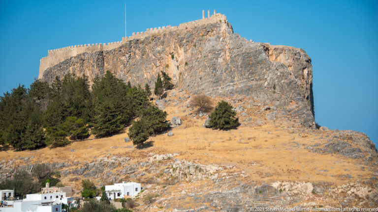 Fortified limestone hill of Lindos Acropolis above a dry slope dotted with pines and scrub under a clear blue sky.