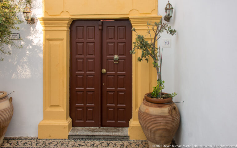 Dark red-brown double door framed by ochre pilasters, pottery jars, plants, and a pebble-mosaic threshold in Lindos.