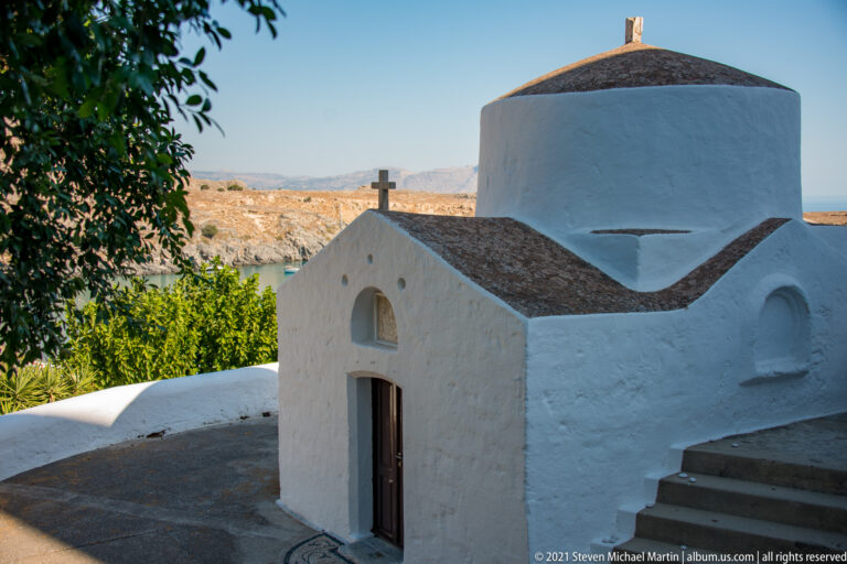Small whitewashed chapel in Lindos with a cross over the entrance and a rounded upper volume against a rocky landscape.