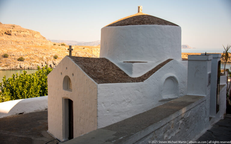 Sunlit whitewashed chapel in Lindos with a cross, rounded upper volume, and dry hills visible beyond.