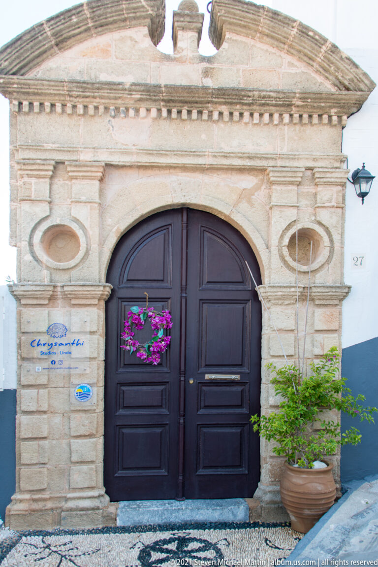 Ornate pale-stone portal with dark double doors, carved pilasters, oval panels, and potted plants in Lindos.