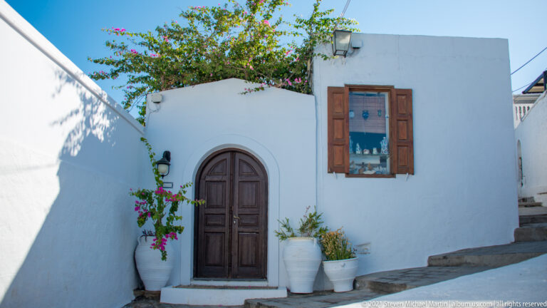 Whitewashed Lindos house with an arched wooden door, shuttered window, potted plants, and bougainvillea beside a sloping lane.