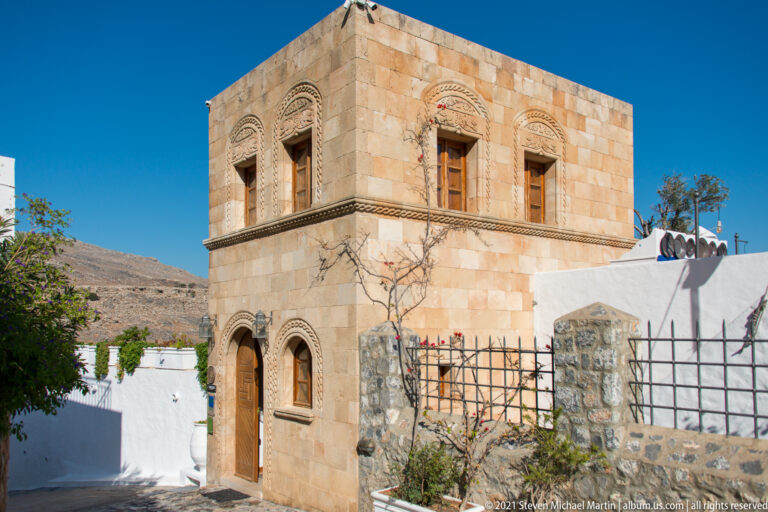 Two-story warm-stone house in Lindos with carved window surrounds, arched lower openings, and decorative horizontal bands.