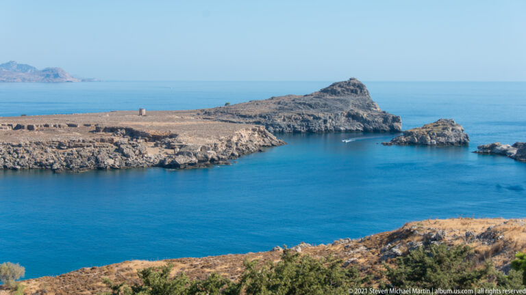 Deep blue Aegean inlet near Lindos framed by pale rocky headlands and small offshore rock formations.