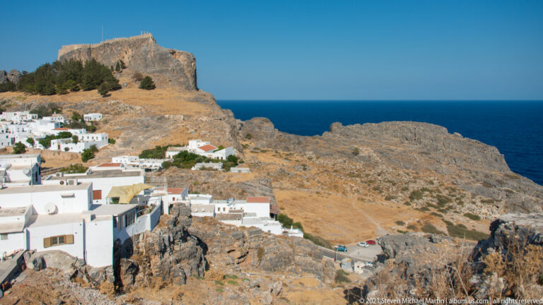 Rocky coastal slope beside whitewashed Lindos houses, descending toward the deep blue Aegean Sea.