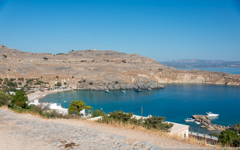 Sheltered blue bay near Lindos with a narrow beach, small harbor, boats, white buildings, and dry rocky hills.
