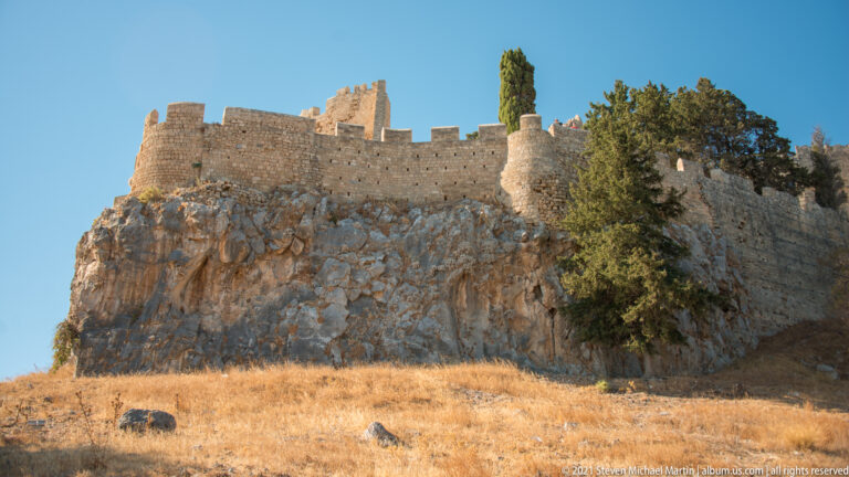 Stone fortification walls and crenellated towers crown a rugged limestone cliff above dry grass and cypress trees.
