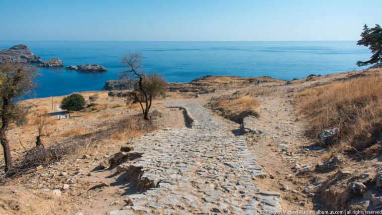 Rough stone-paved path descends through a dry coastal slope toward the blue Aegean Sea and offshore rocks.