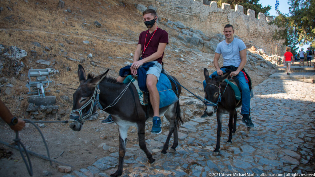 Two visitors ride saddled donkeys up a cobbled path beside the stone walls of Lindos Acropolis.