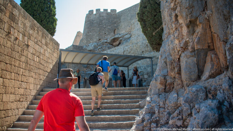 Visitors climb a narrow stone stairway between masonry walls and exposed rock toward the fortified Lindos Acropolis.