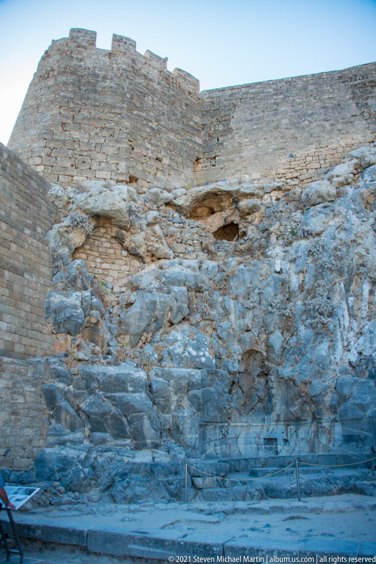 High stone wall with crenellations rises directly from exposed limestone bedrock inside Lindos Acropolis.