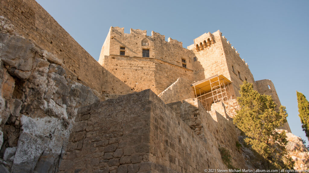Massive stone walls and crenellated towers rise above the entrance approach, with conservation scaffolding on one section.