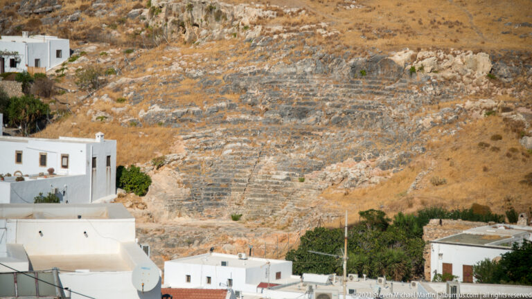 Exposed banded limestone hillside above whitewashed buildings in Lindos, with dry grass and sparse vegetation.