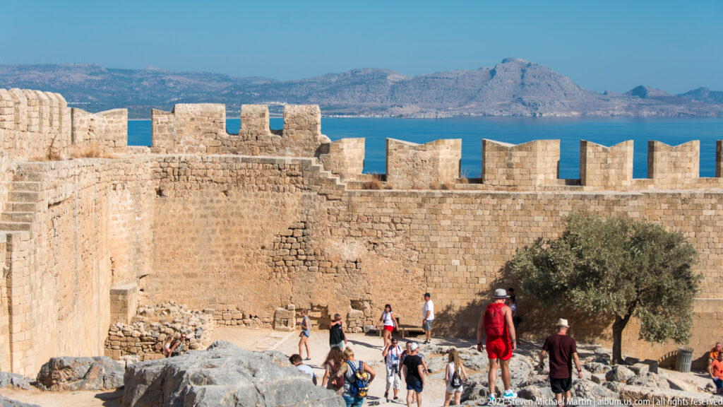 Crenellated stone wall of Lindos Acropolis with visitors, blue sea and distant mountainous coast beyond.