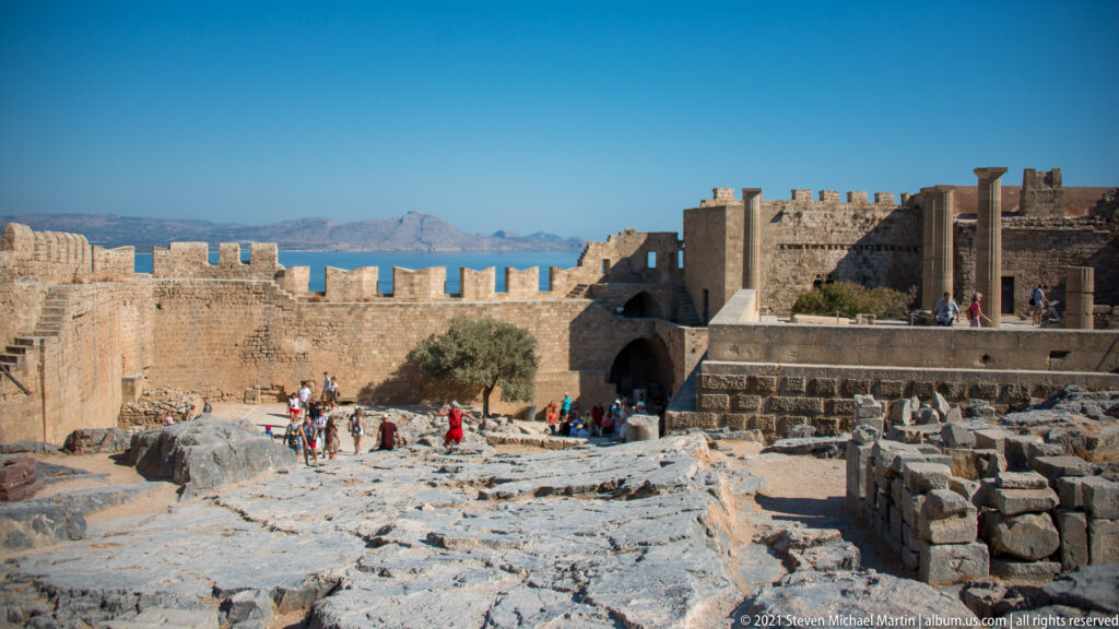 Wide interior view of Lindos Acropolis with bedrock, ruined foundations, standing columns, visitors and crenellated walls.