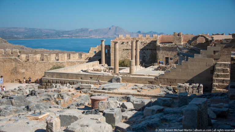 Elevated view across rocky foundations, terraces and standing columns inside Lindos Acropolis with sea beyond.