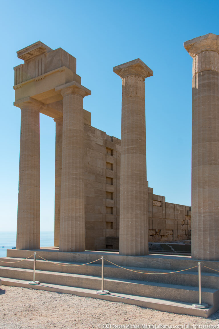 Restored stone columns and lintels stand before fragmentary walls, with the Aegean Sea visible through the openings.