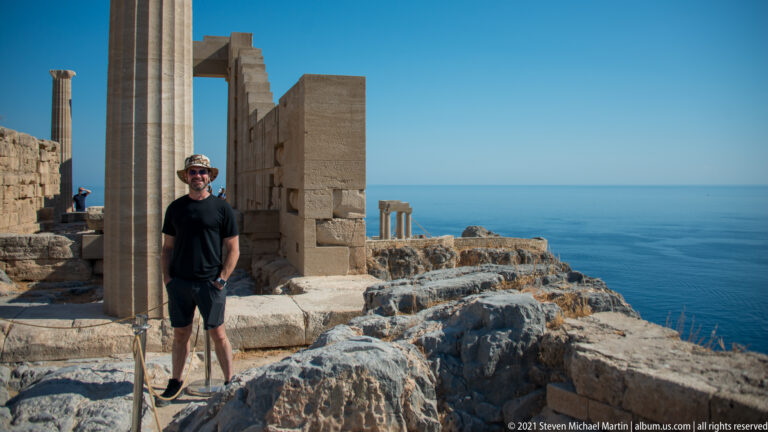 Visitor stands beside tall restored columns on rocky ground, with the Aegean Sea and distant ruins behind.