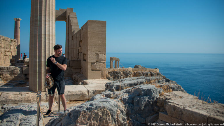 Visitor gestures beside a restored stone column, with block walls, limestone bedrock and blue sea beyond.