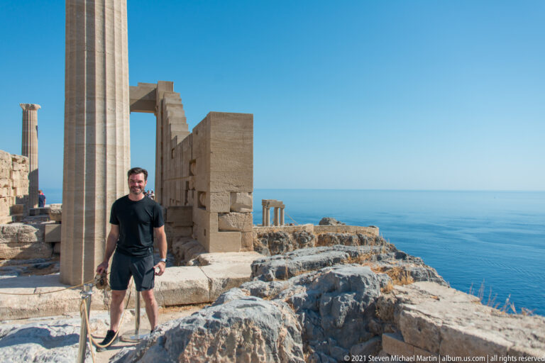 Visitor stands between restored columns and stone walls on the Lindos Acropolis, with the Aegean Sea behind.