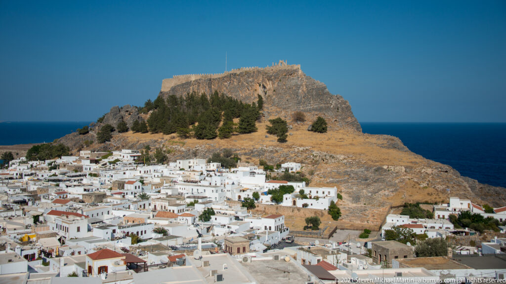 Dense whitewashed village of Lindos spread below the fortified acropolis, with pines on the slope and sea behind.