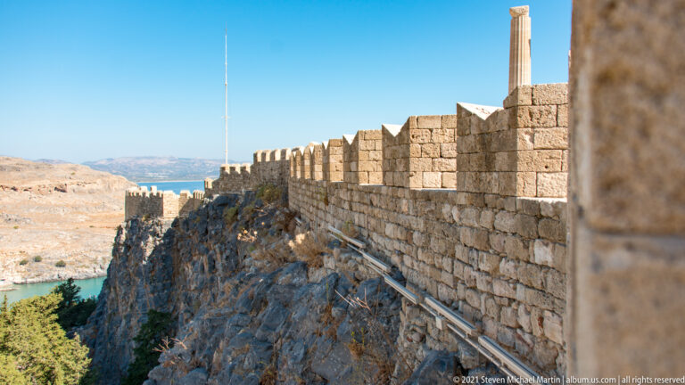 Stone crenellated wall follows the cliff edge of Lindos Acropolis above a steep rocky drop.