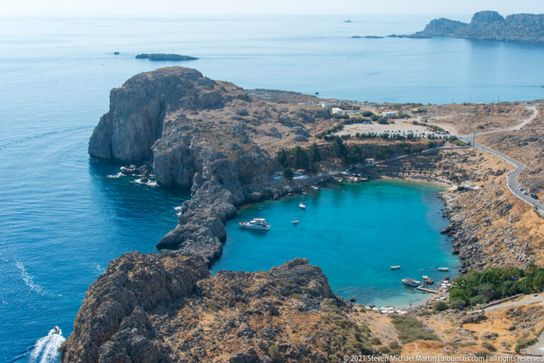 Turquoise St Paul’s Bay lies within a rocky headland below Lindos, with small boats and the open sea beyond.