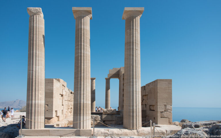 Three tall restored columns rise before fragmentary sanctuary walls, with blue sky and sea visible through the ruins.
