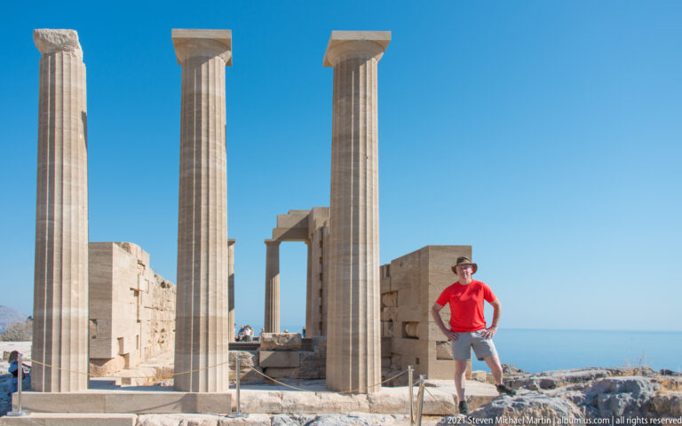 Visitor in red stands between tall restored columns on Lindos Acropolis, with masonry ruins and sea behind.