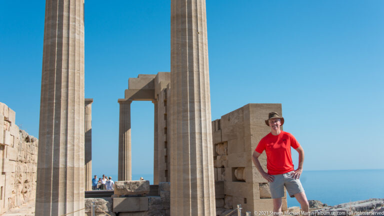 Visitor in red stands beside towering restored columns, with block masonry, an architectural opening and sea behind.