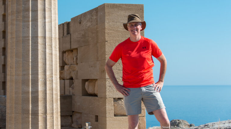 Visitor in red stands beside a massive stone pier and restored fluted column, with the Aegean Sea beyond.