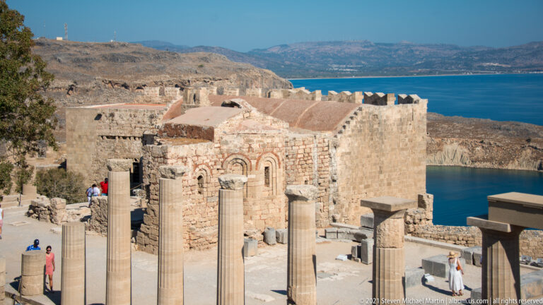 Weathered stone Church of St John rises behind ancient columns inside Lindos Acropolis, with the blue sea beyond.