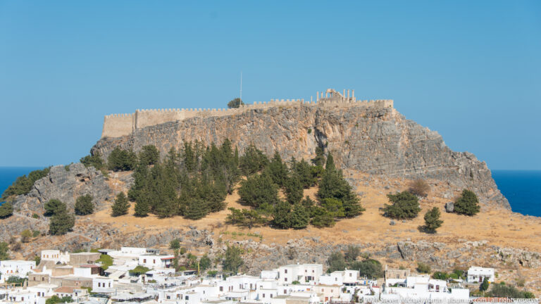 Lindos Acropolis rises above white village rooftops, with defensive walls on the rocky summit and pines below.
