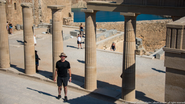 Visitor in dark clothing stands beneath tall restored columns, with other visitors and stone walls visible beyond.