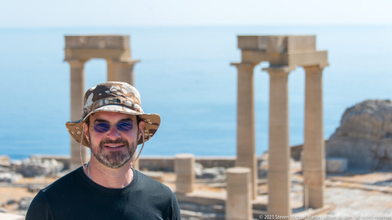 Visitor in hat and sunglasses stands before restored stone columns with the blue Aegean Sea beyond.