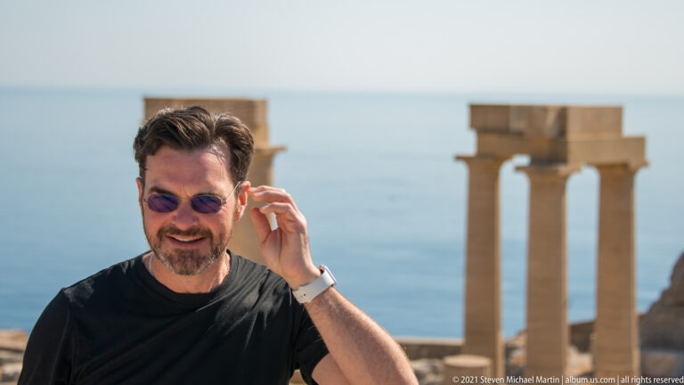 Visitor adjusts sunglasses in front of restored stone columns and the blue sea at Lindos Acropolis.