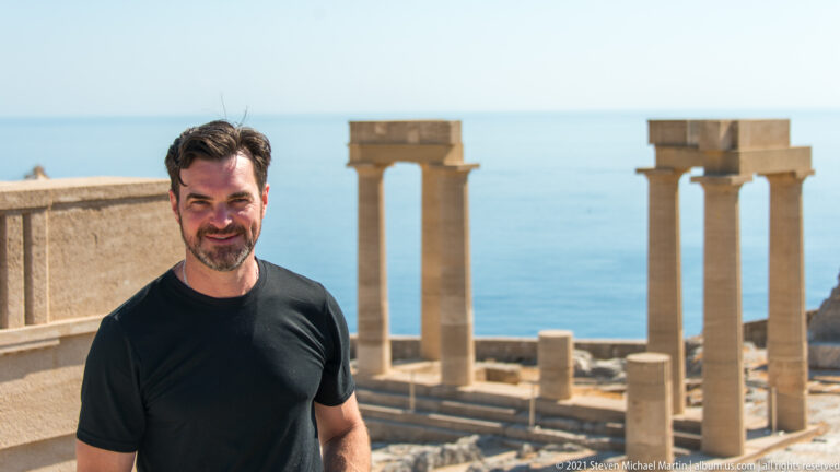 Visitor stands between restored stone columns with masonry ruins and the Aegean Sea behind.