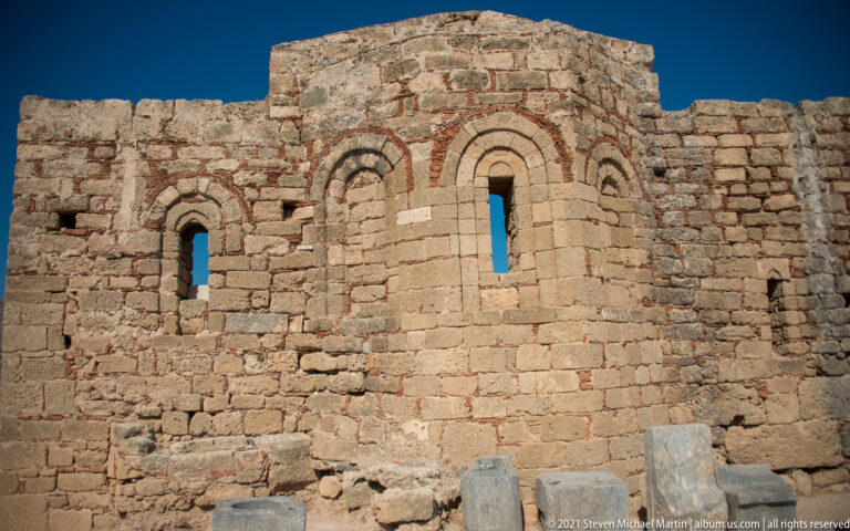 Stone church wall with narrow arched windows, brick accents and architectural blocks at Lindos Acropolis.