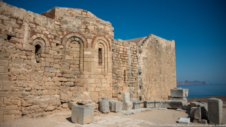 Weathered stone church with arched windows stands among carved blocks and ruins, with the sea beyond.