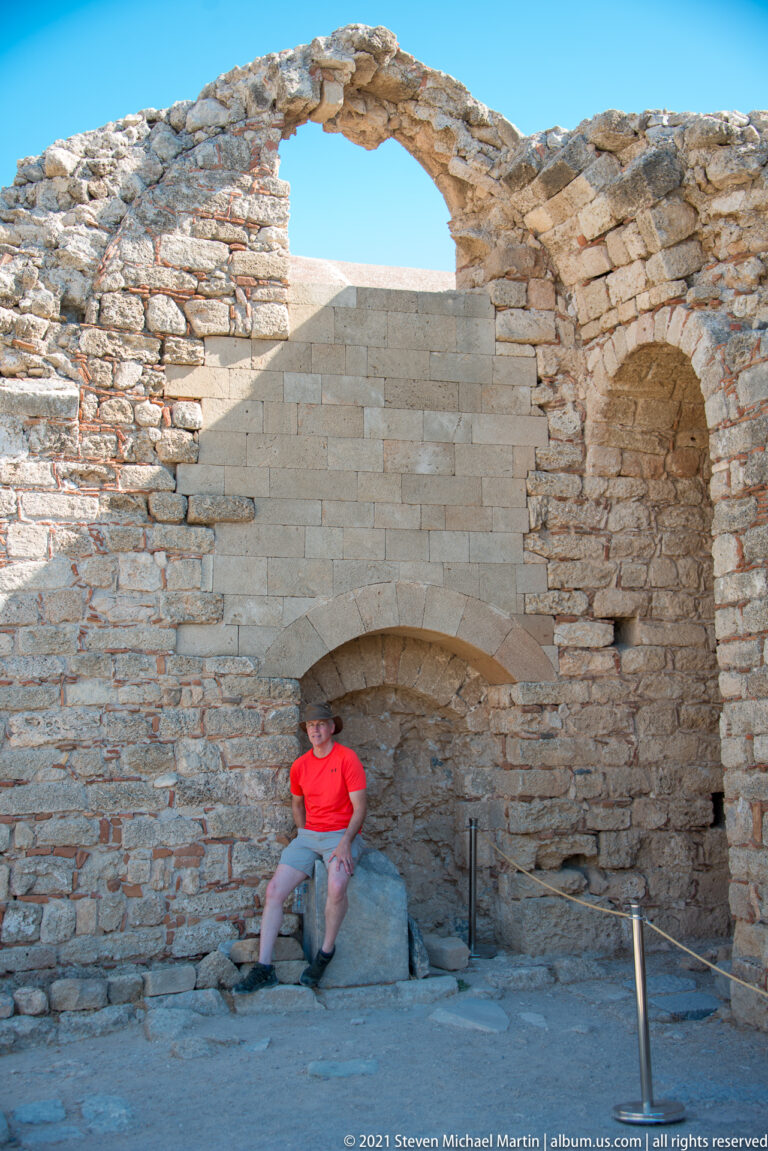Visitor sits inside a roofless stone church with vaulted recesses and rough masonry at Lindos Acropolis.