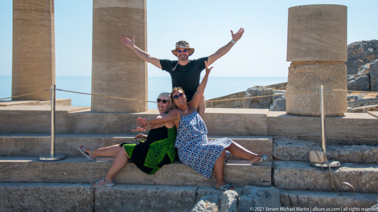 Three visitors pose on stone steps between restored columns with the blue Aegean Sea behind.