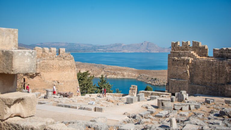 Ruined foundations and blocks lie below crenellated fortifications, with blue coastal water and mountains beyond.