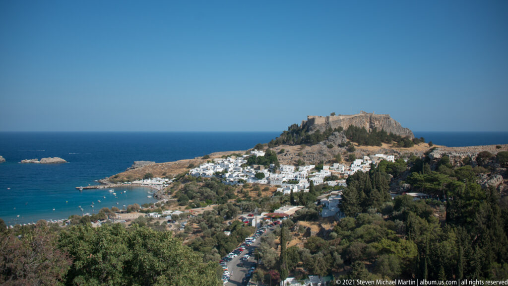 Distant Lindos village and fortified acropolis above a sheltered blue bay, seen across trees and a winding road.