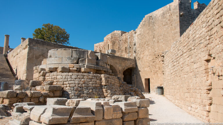 Rounded stone foundation and scattered architectural blocks sit below high fortress walls inside Lindos Acropolis.