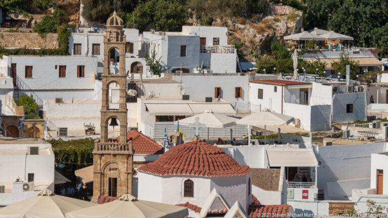 Stone bell tower and red-tiled church roof rise among whitewashed houses and terraces in Lindos village.