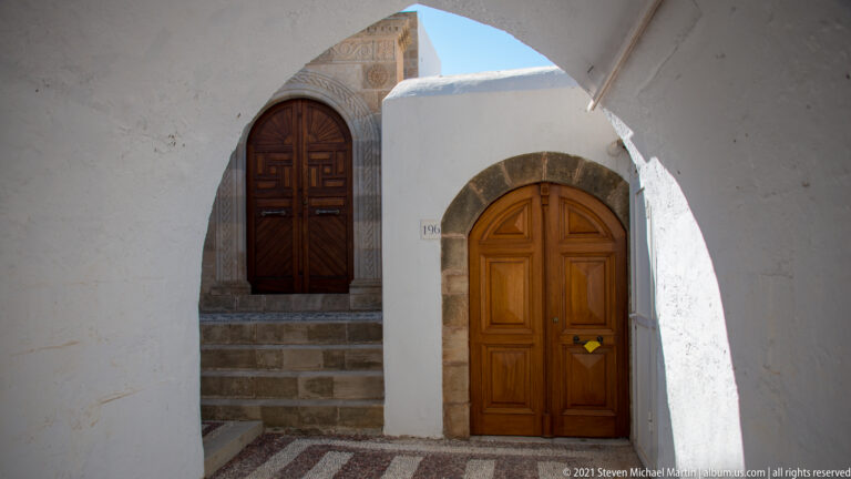 Two wooden doors in a whitewashed passage, one above stone steps and one beneath a stone arch.