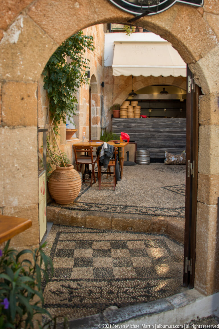 Stone arch frames a shaded courtyard with black-and-white pebble paving, tables, plants and a closed counter.
