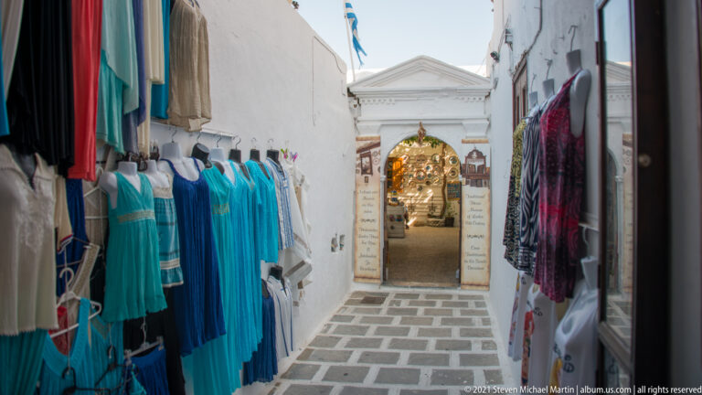 Narrow whitewashed shopping passage lined with turquoise and patterned clothing leading to a small illuminated shop.