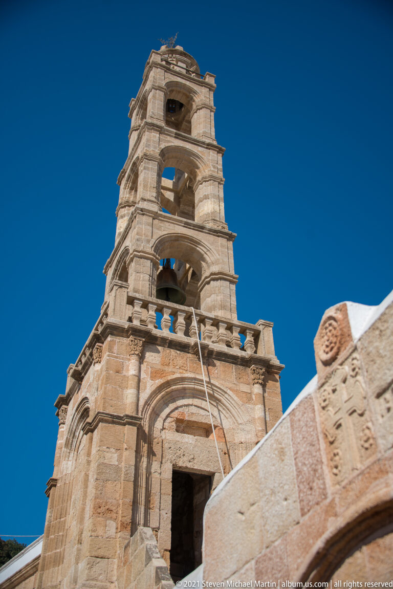Ornate multi-tiered stone bell tower with arched openings rises against a deep blue sky in Lindos.
