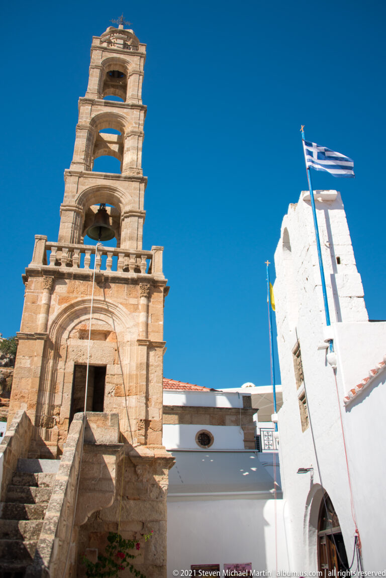 Stone bell tower rises beside whitewashed church walls and a Greek flag above a flight of steps.